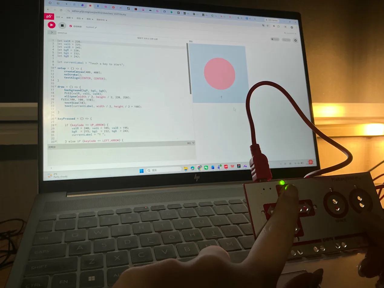 Hand holds Makey Makey up to a laptop running a pink circle on a blue P5.js canvas.