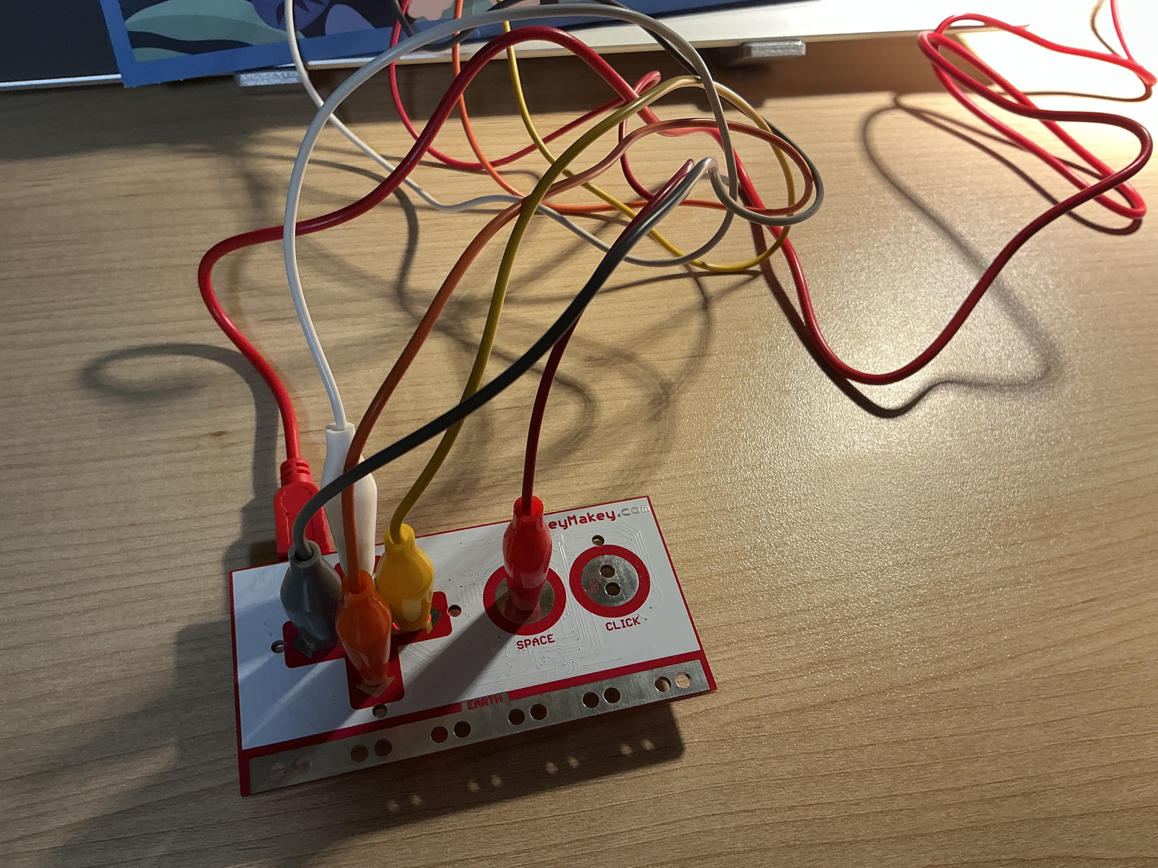 The image displays the Makey Makey board placed on the desk with five colored alligator clip wires fanning outward, showing the full wiring layout before connecting to the physical interface.
