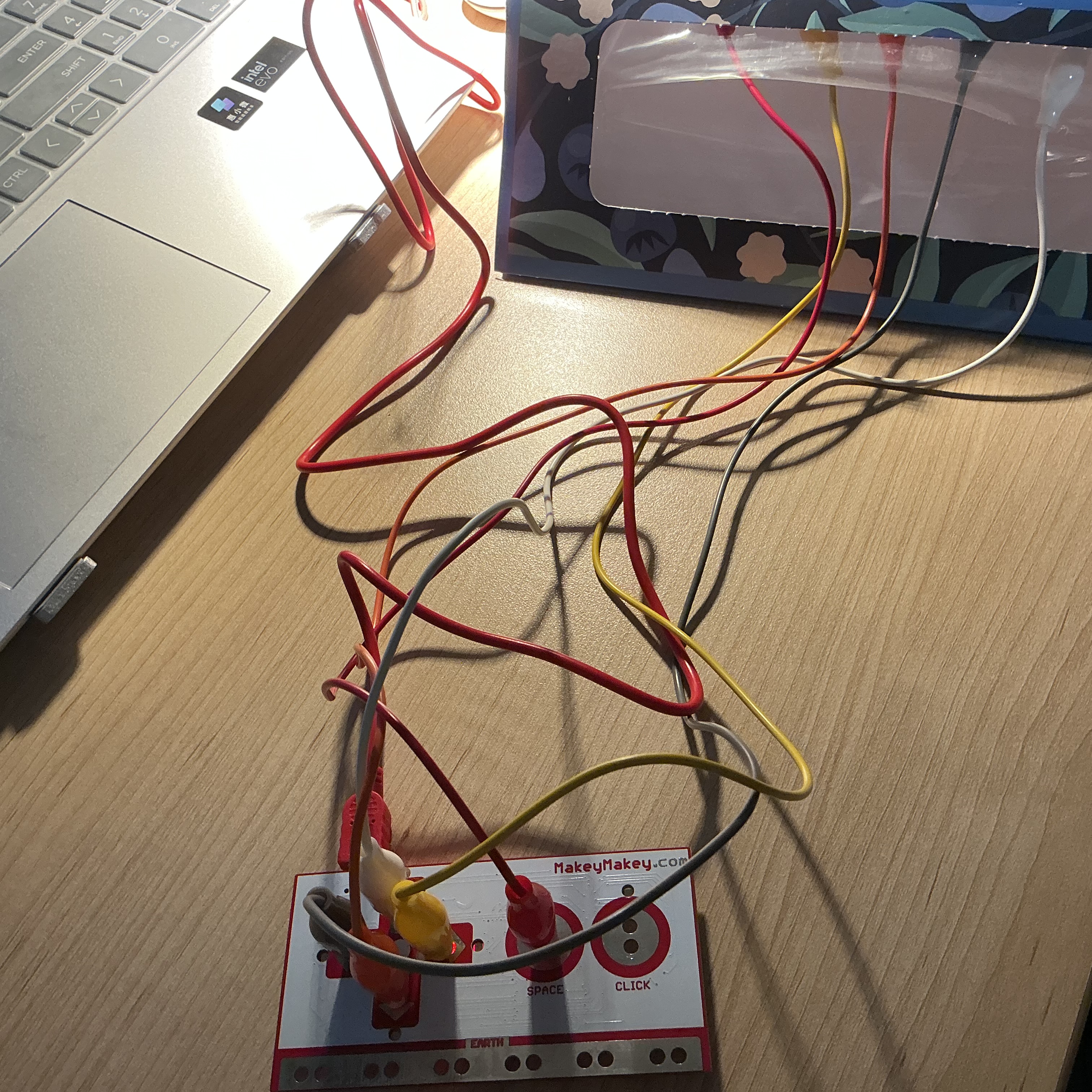 The image displays a laptop with tangled colored wires leading to the blue floral box partially visible in frame, showing the wiring in progress during setup of the magnet and copper tape system.