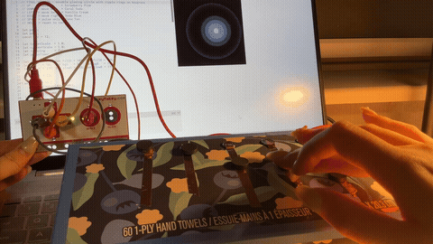 The image displays a hand pressing the magnet-topped copper tape buttons on a camouflage-patterned box while a warm orange glow pulses on the laptop screen, using copper tape and magnets as the combined conductive trigger system.