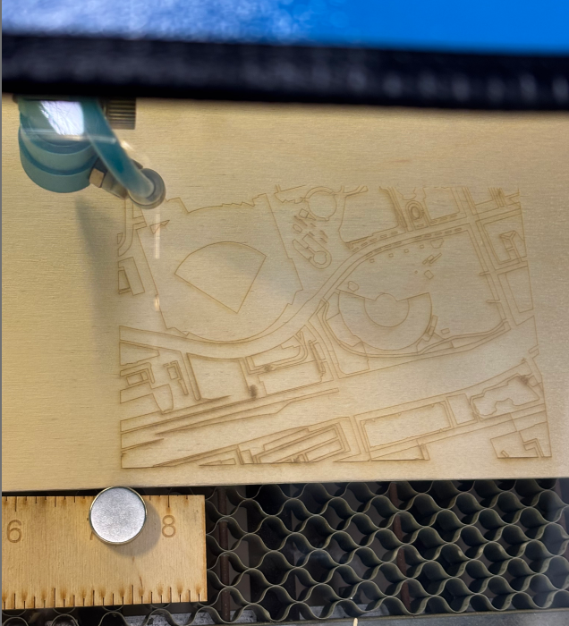 The image depicts the laser-cutting process of a wooden map, with the tool working on intricate details.